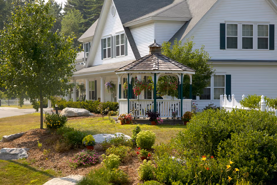 A white two-story house with green shutters surrounded by a well-maintained garden. In front of the house is a white gazebo with a shingled roof, decorated with hanging flower baskets. The garden features various shrubs, flowers, and small trees, with a white picket fence visible on the right side.
