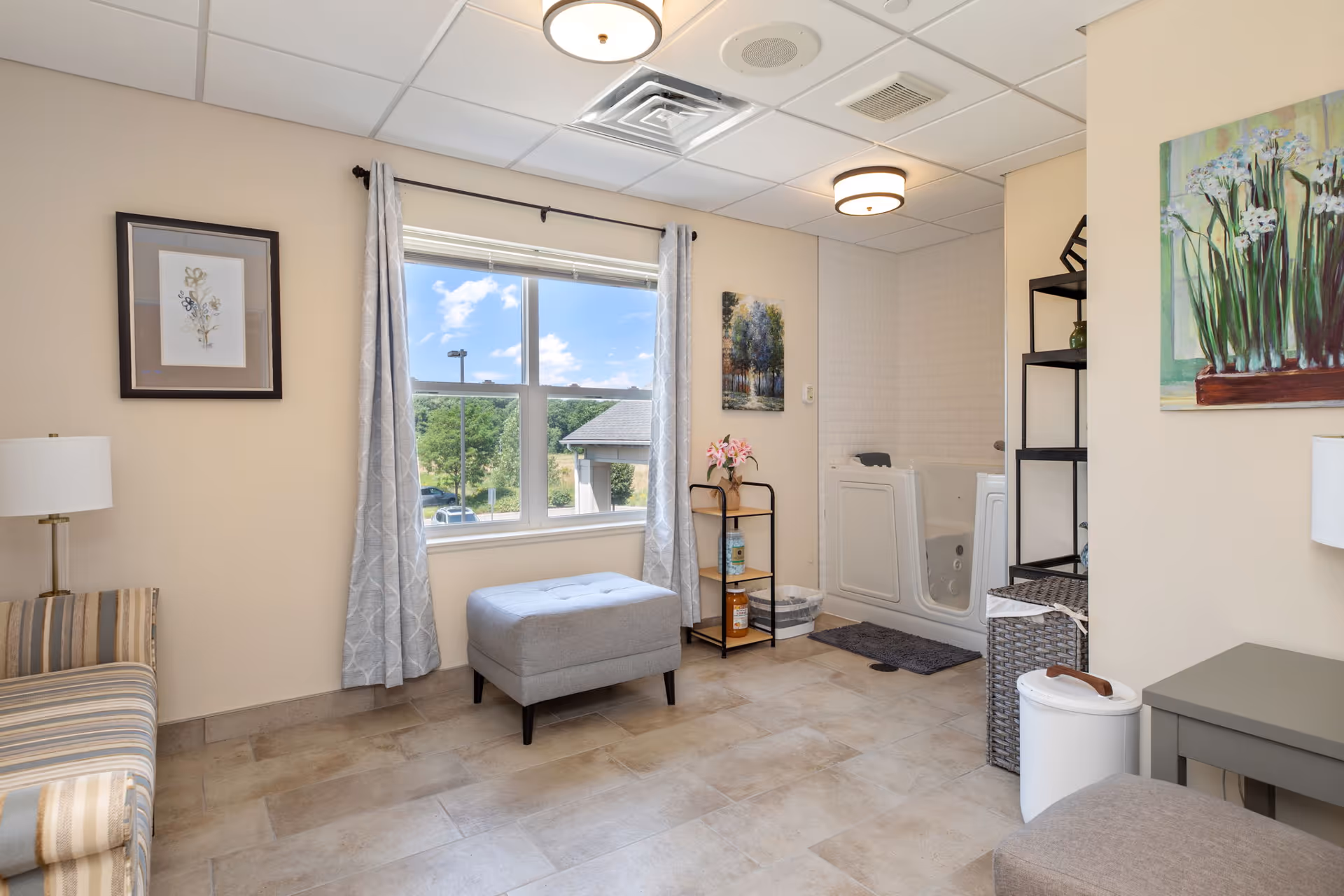A bright room with beige walls and tiled floor featuring a striped sofa on the left, a gray ottoman in front of a window with light gray curtains, and a walk-in bathtub in the corner. The room is decorated with framed artwork on the walls, a small black metal shelf with flowers and toiletries, a gray side table, and a white trash bin. Ceiling lights illuminate the space.