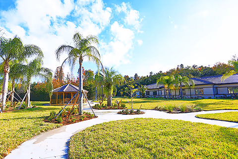 A sunny outdoor garden area with a curved concrete pathway, green grass, palm trees, and a small gazebo. A single-story building is visible in the background under a partly cloudy blue sky.