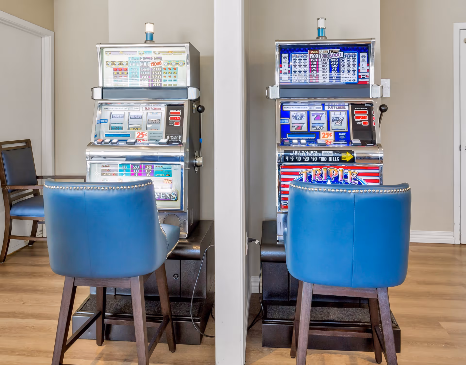 Two vintage slot machines placed side by side with a partition wall between them. Each slot machine has a blue cushioned chair in front of it. The floor is wooden, and there is an additional wooden chair with a dark cushion to the left side of the left slot machine. The background walls are light-colored.