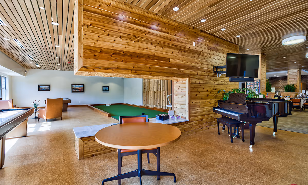 Open wood-paneled senior living common room with a grand piano, round table, seating and an indoor putting green.