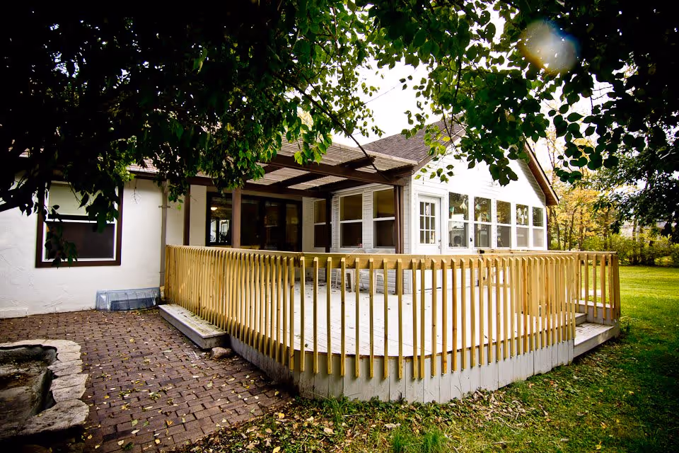 Exterior view of a single-story home with a wooden deck surrounded by a wooden railing. The house has white siding and multiple windows, with a covered porch area. The yard is grassy with trees providing shade.