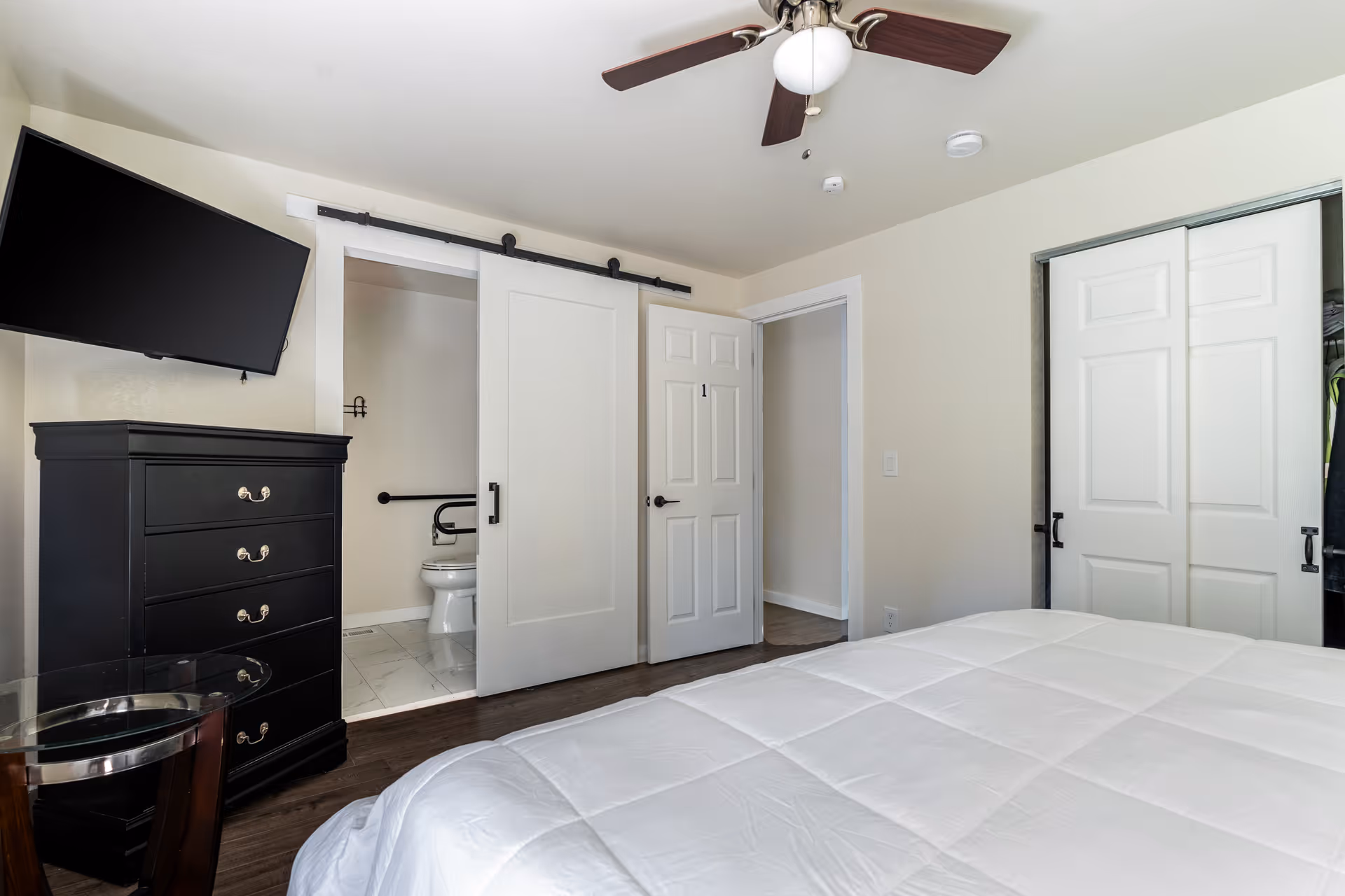 Clean bedroom with a bed in the foreground, wall-mounted TV and dresser, a sliding barn door open to a bathroom, and closet doors.