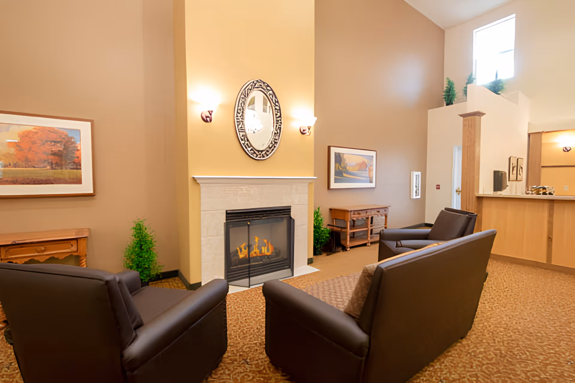 A cozy senior living lounge with leather chairs arranged around a lit fireplace, framed artwork, and a reception desk area.