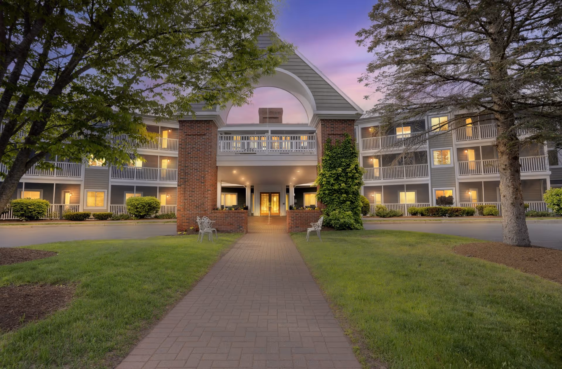 Front entrance of a three-story senior living building with balconies, a covered porte-cochère and a paved walkway through a lawn at dusk.