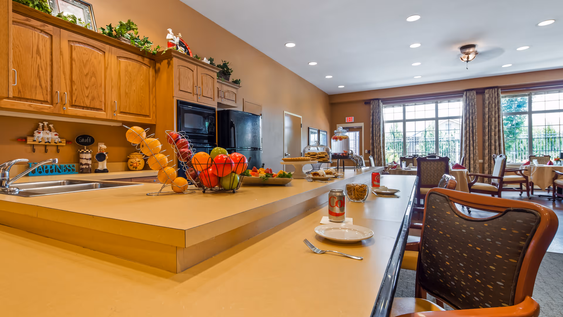 A bright and spacious dining area in an assisted living facility featuring a long counter with chairs, plates, cutlery, and cans of Diet Coke. The kitchen area has wooden cabinets, a black refrigerator, and a microwave. The counter displays decorative fruit baskets and baked goods. Large windows with curtains allow natural light to fill the room.
