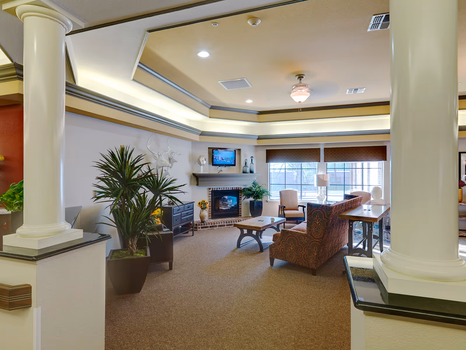 Bright furnished common living room with couches and armchairs around a fireplace and wall-mounted TV, large windows, decorative columns, and potted plants.