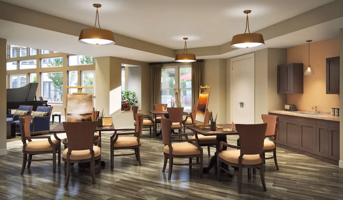Sunlit communal activity/dining room with round tables and chairs, art easels, a kitchenette, and lounge seating by large windows.