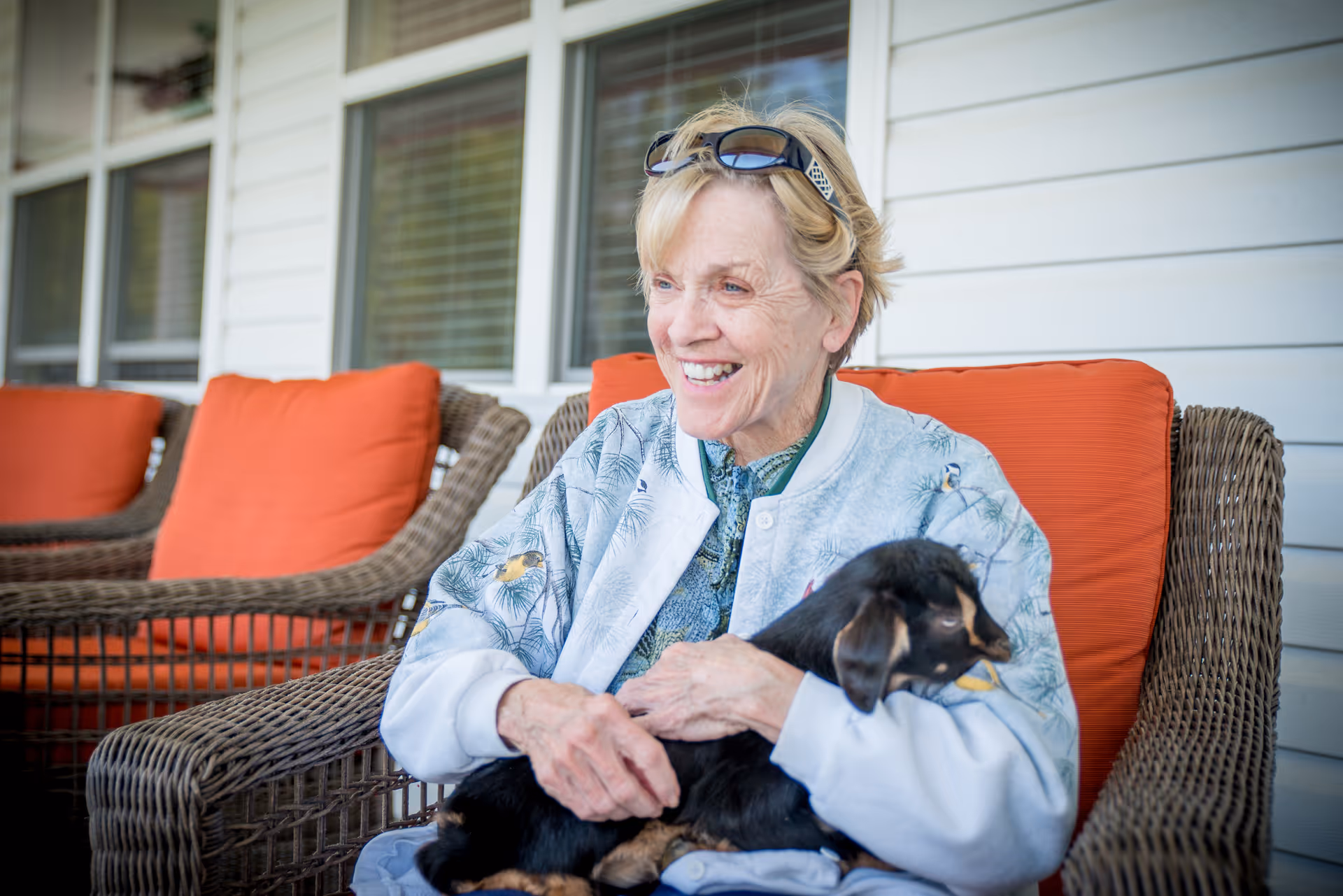 An elderly woman sitting on a wicker chair with orange cushions on a porch, smiling and holding a small black and brown goat in her lap. She is wearing a light-colored jacket with bird patterns and sunglasses on her head.