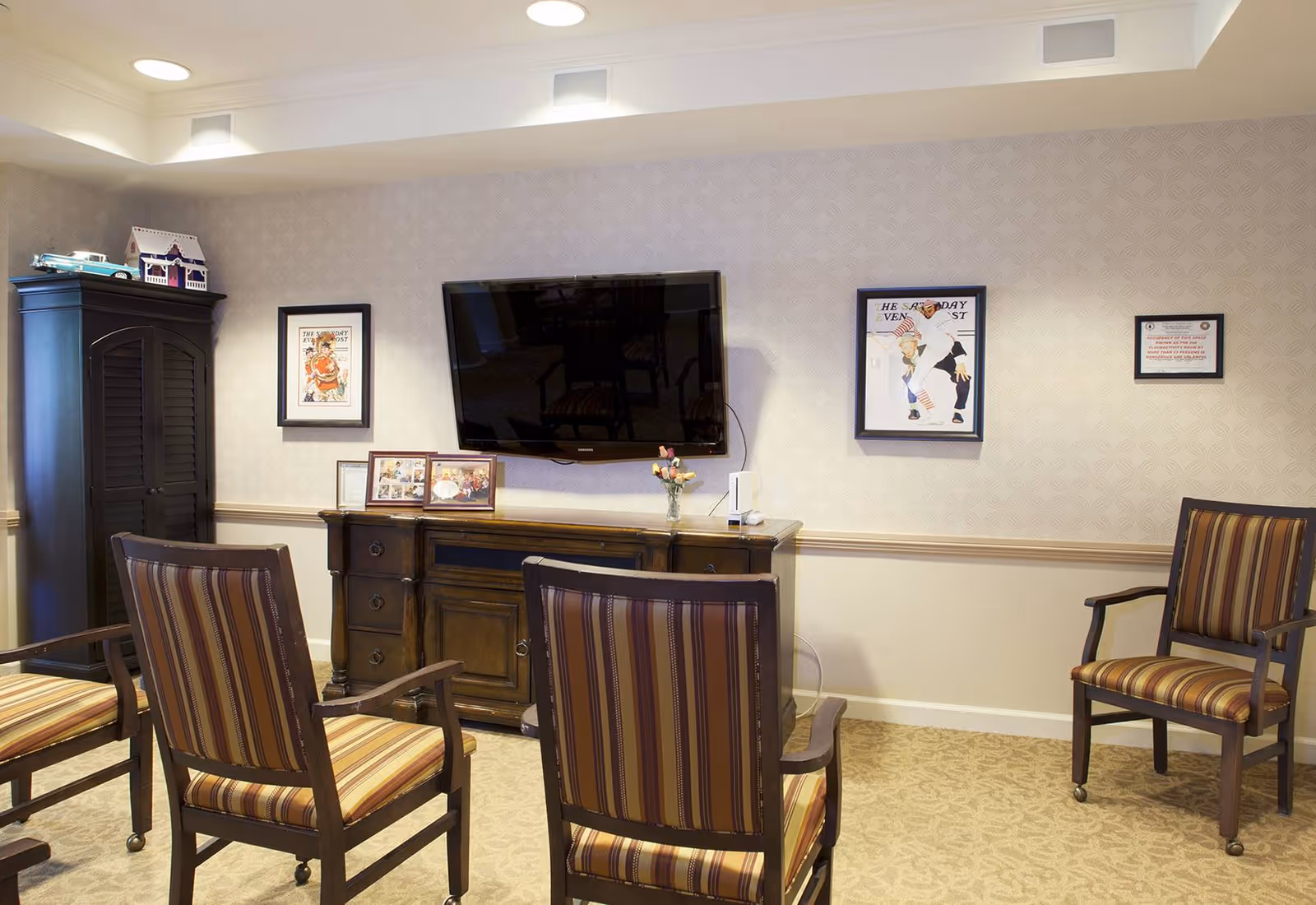 A small common room with striped wooden armchairs arranged facing a wall-mounted flat-screen TV above a wooden console and framed pictures.