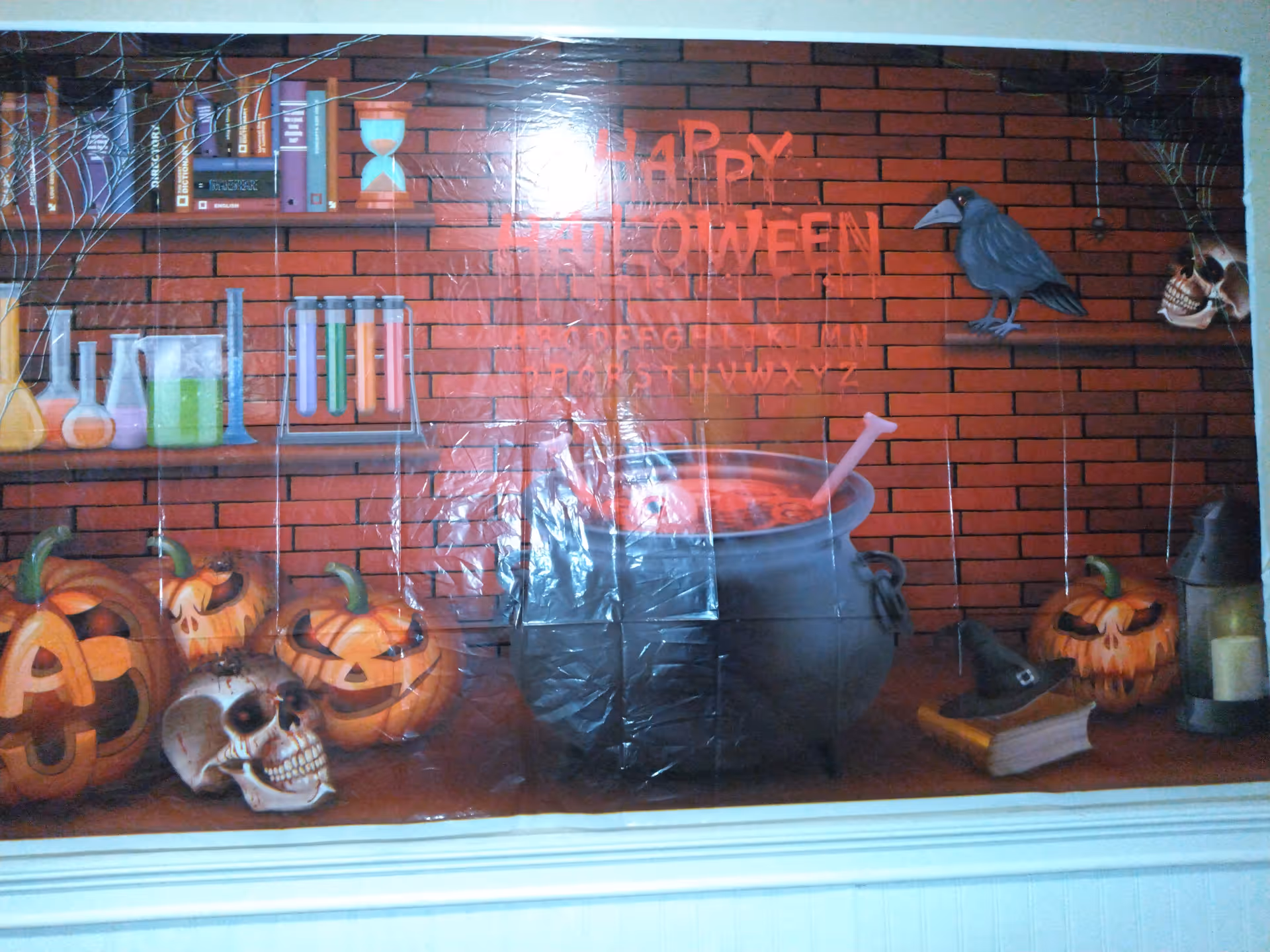 A Halloween-themed wall decoration featuring a brick wall background with shelves holding books, test tubes, and beakers. Below the shelves are carved pumpkins with spooky faces, a skull, a large black cauldron with a stirring spoon, a black crow perched on a shelf, a witch's hat on a book, and a lantern with a candle. The words 'Happy Halloween' are written in red on the brick wall.