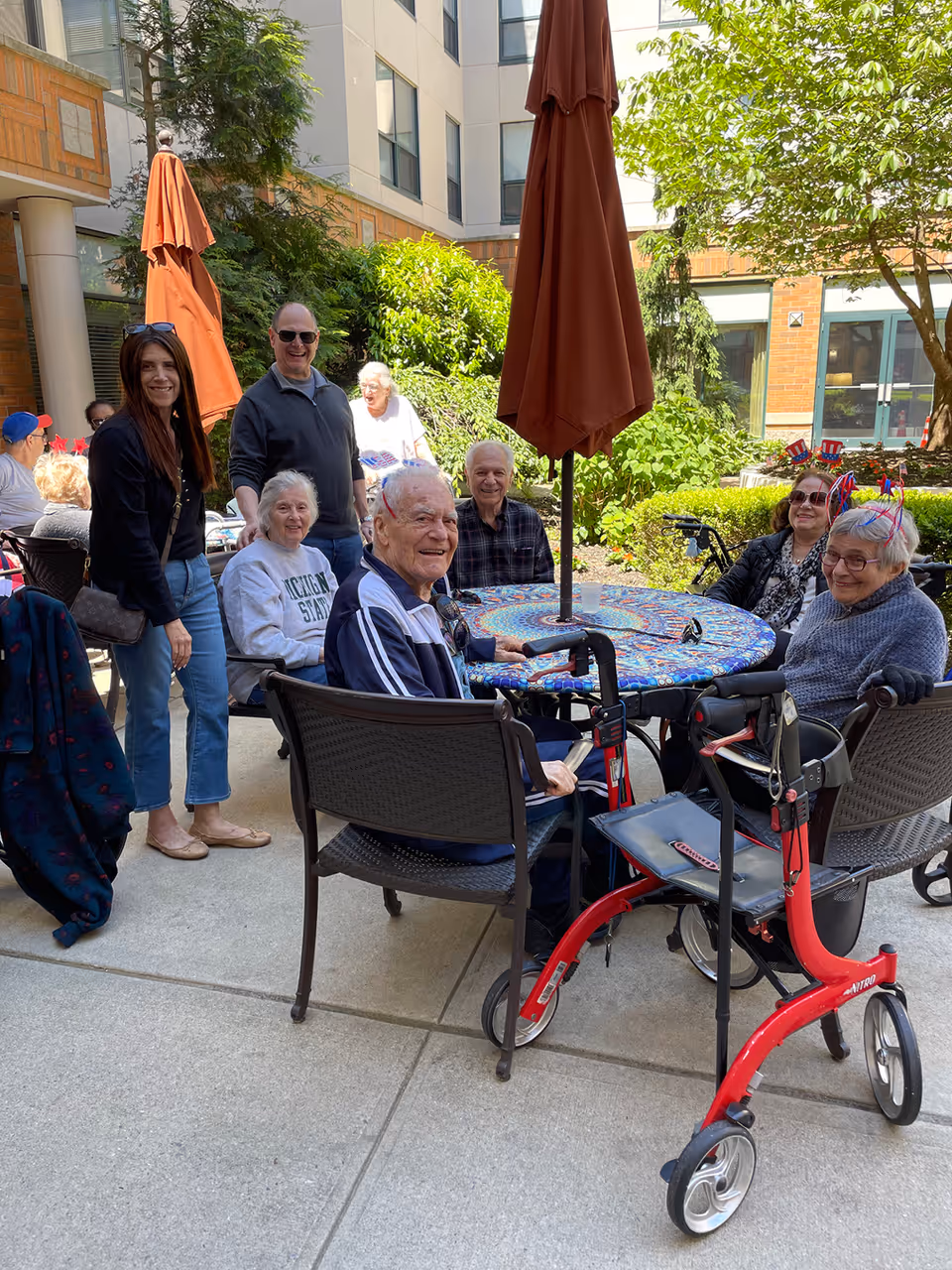 A group of elderly people and two adults gathered around a round outdoor table with a colorful tablecloth and a closed umbrella in the center. They are sitting on patio chairs in a courtyard area with greenery and a building in the background. One elderly woman is wearing festive headbands, and a red walker is visible in the foreground.