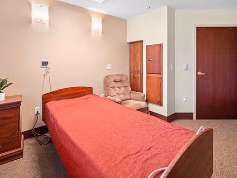 A simple bedroom in a senior living facility with a single bed covered in a red blanket, a cushioned armchair, a wooden nightstand with a small plant, beige walls with two wall-mounted lights, and a closed wooden door.
