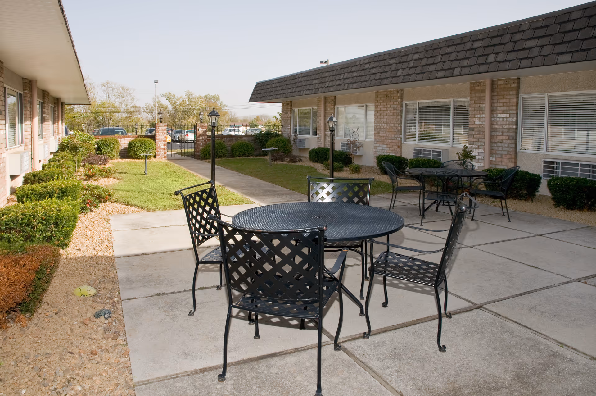 Outdoor patio area at Life Care Center of Morristown with black metal tables and chairs on a concrete surface, surrounded by bushes and grass, with a building featuring windows and brick accents in the background.