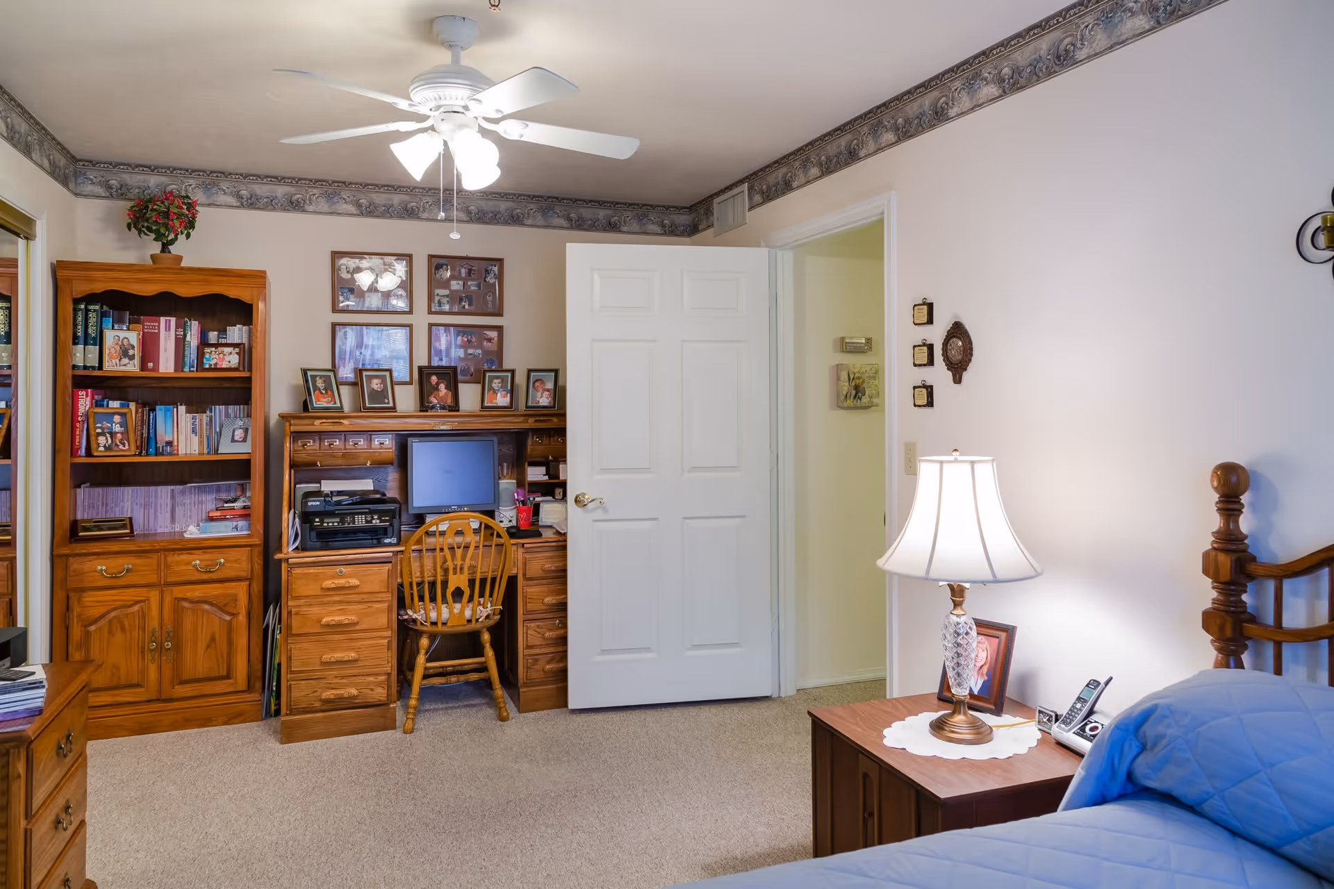 A cozy bedroom with a wooden bed partially visible on the right, covered with a blue quilt. Next to the bed is a wooden nightstand with a decorative lamp, a framed photo, and a cordless phone. Across the room is a wooden desk with a computer monitor, printer, and several framed photos on top. To the left of the desk is a wooden bookshelf filled with books and more framed photos. The room has beige carpet, white walls with a decorative border near the ceiling, and a white ceiling fan with lights. The door to the room is open, revealing a hallway.