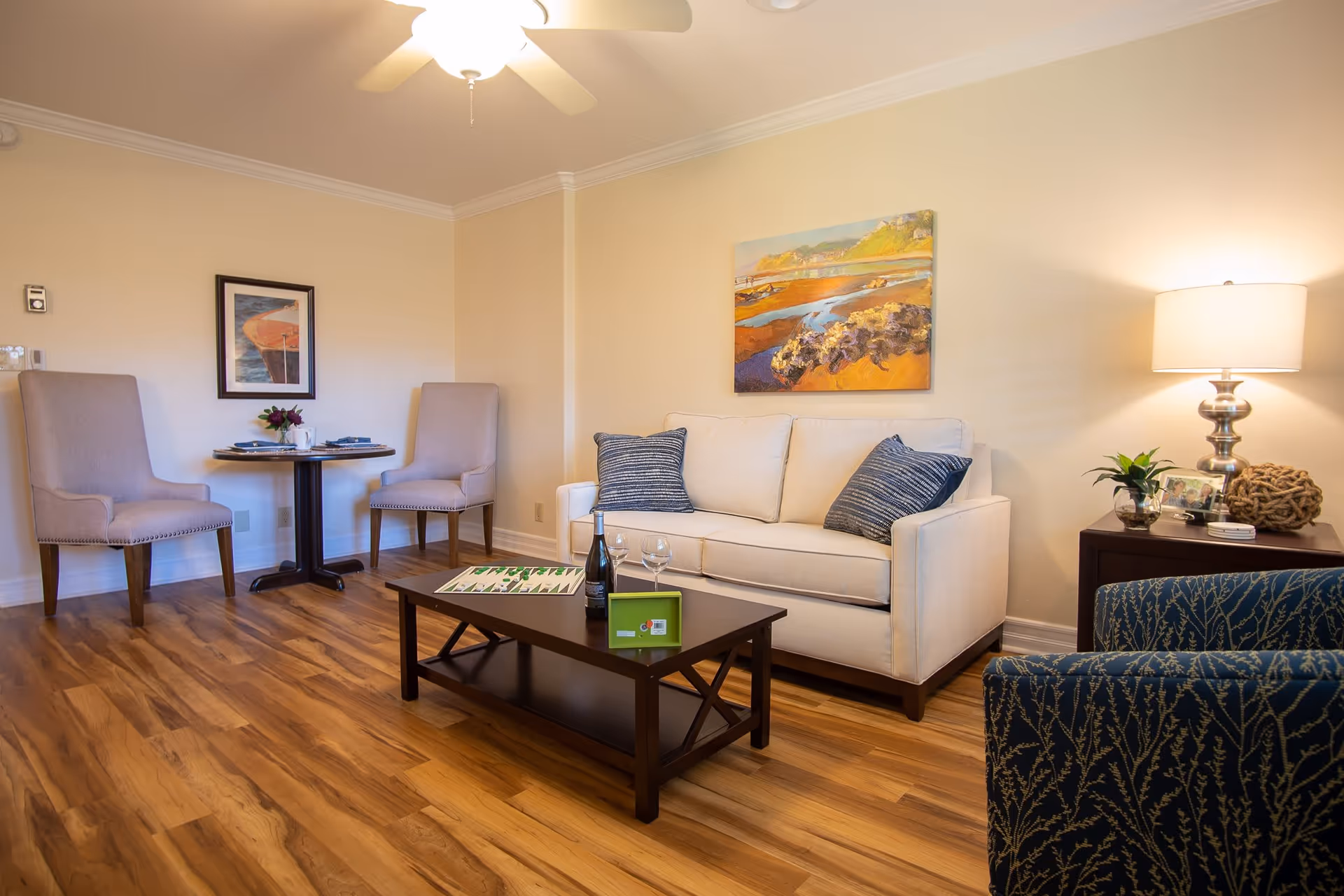 A cozy living room with a beige sofa adorned with two blue patterned pillows, a dark wooden coffee table with a bottle of wine, two wine glasses, and a game board on it. To the left, there is a small round table set for two with beige upholstered chairs. A blue patterned armchair is partially visible on the right. The room has wooden flooring, a ceiling fan with a light, a table lamp on a side table, and two framed artworks on the walls.