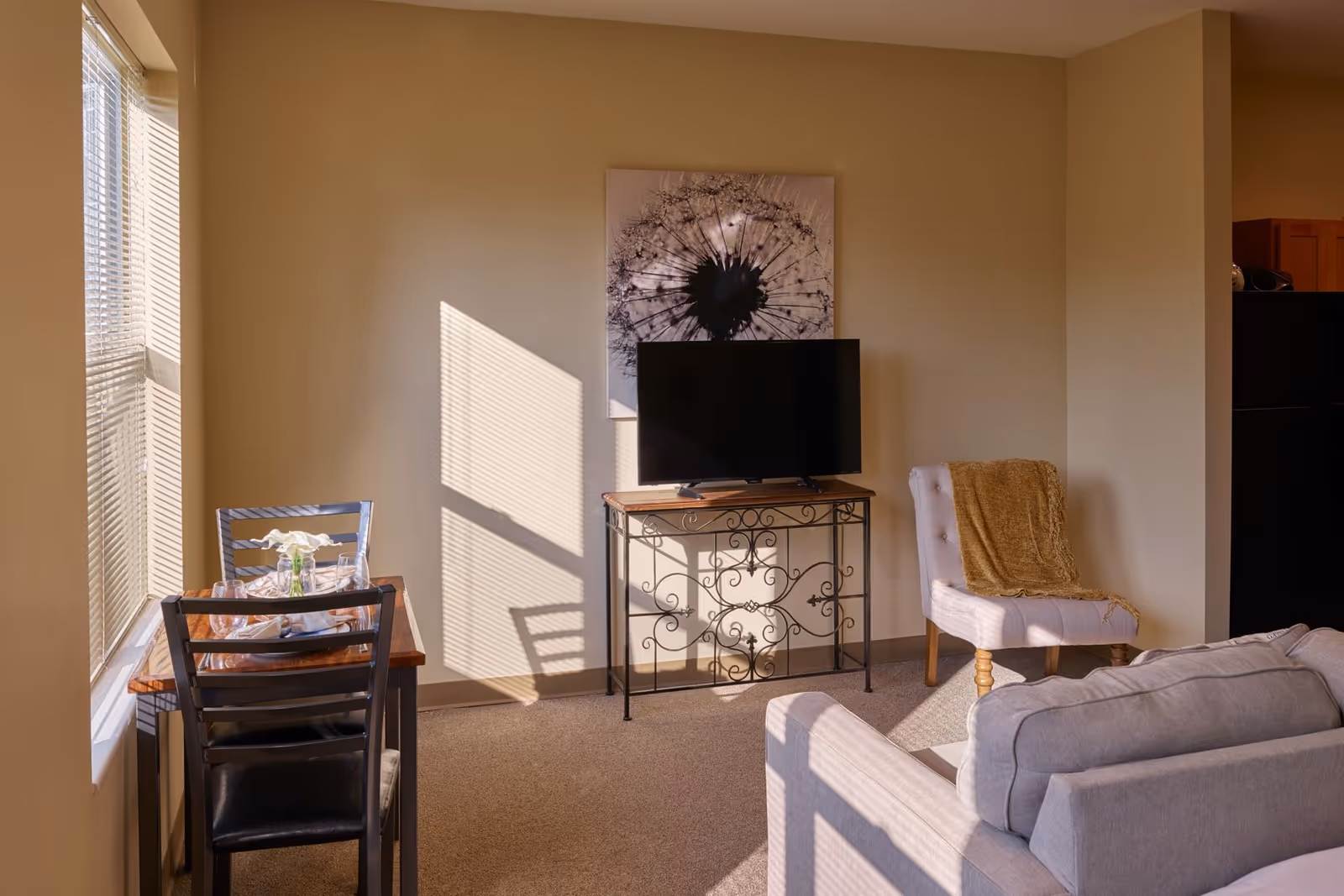 A cozy living room area with a small dining table set for two near a window with blinds. A flat-screen TV sits on a decorative metal and wood stand against a beige wall with a large dandelion artwork above it. There is a cushioned chair with a brown throw blanket and part of a light gray sofa visible in the foreground.