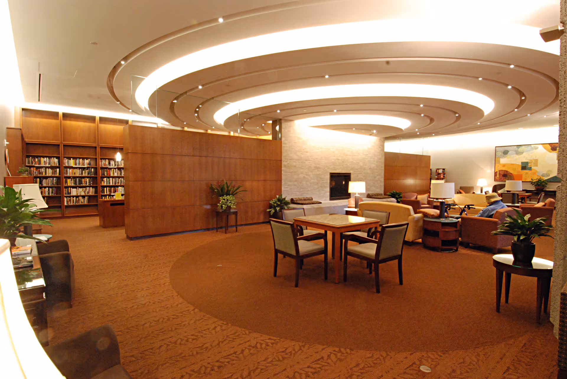 A spacious, well-lit common area with a circular ceiling design featuring concentric light rings. The room includes a fireplace with a stone surround, several seating areas with armchairs and sofas, a table with four chairs, potted plants, and a bookshelf filled with books in the background. A person wearing a hat is seated on one of the sofas.