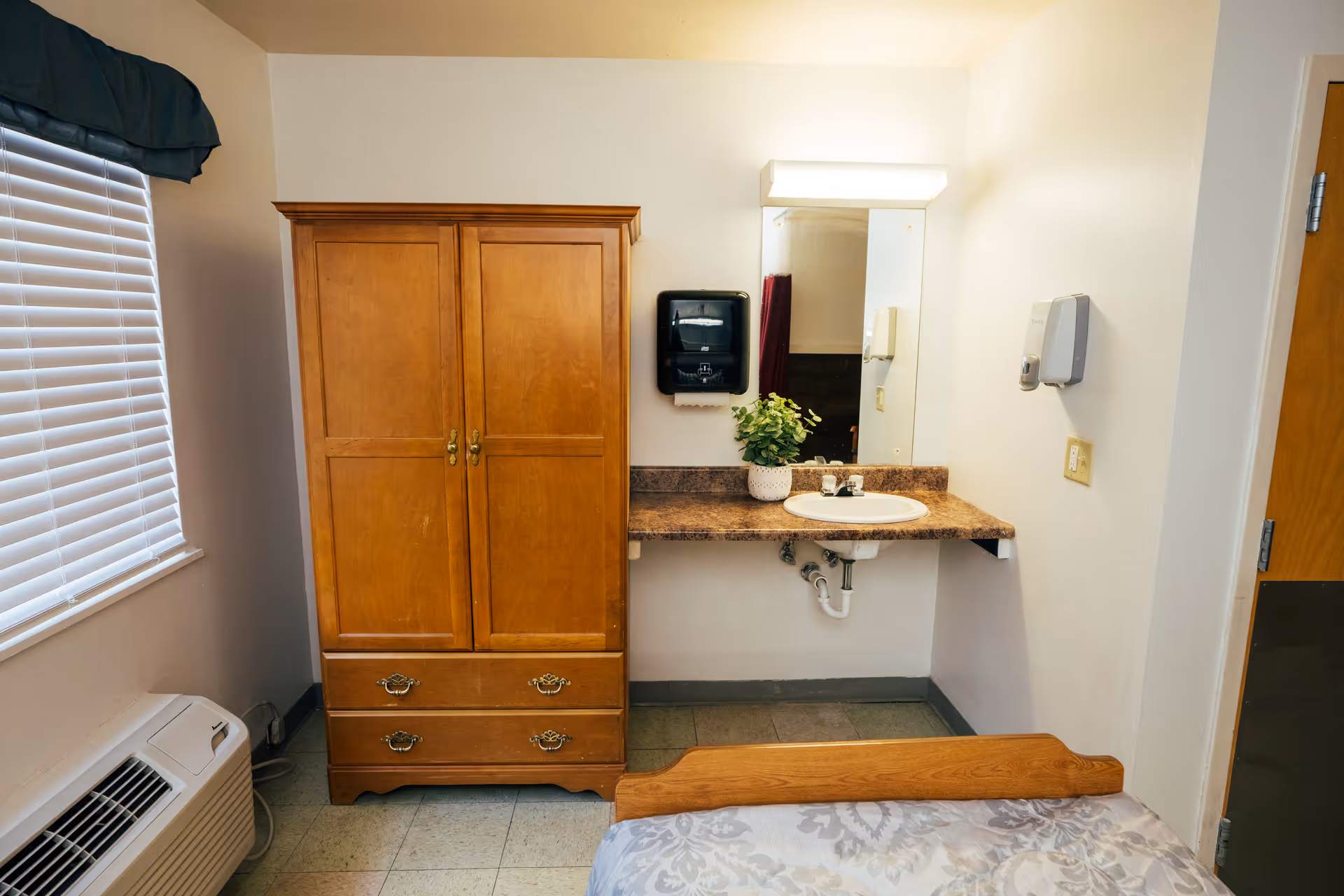 A small room with a wooden wardrobe, a bed with a patterned bedspread, a wall-mounted sink with a mirror above it, a paper towel dispenser, a soap dispenser, and a window with closed blinds and a dark valance.