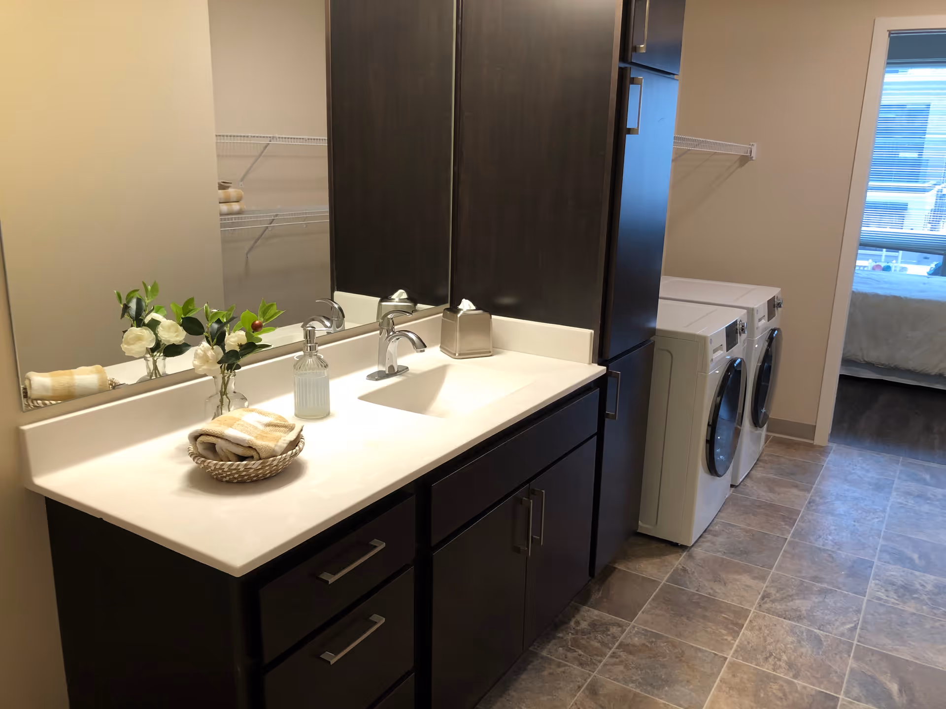 A bathroom vanity with sink, mirror and dark cabinets next to side-by-side washer and dryer in a tiled interior leading to a bedroom.