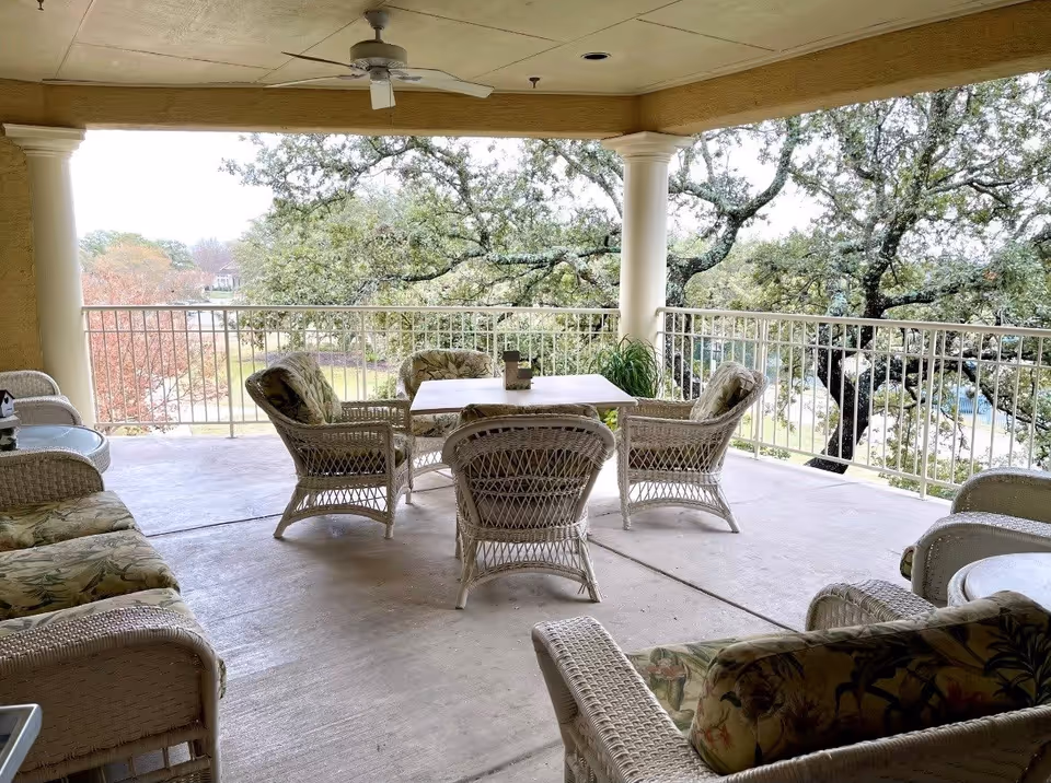 Covered outdoor patio area with wicker furniture including cushioned chairs and a table, overlooking trees and a landscaped area beyond a white railing.
