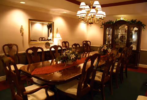 A formal dining room with a long wooden table surrounded by ten cushioned chairs. The table is decorated with a red runner and floral centerpieces. A chandelier with multiple lampshades hangs above the table. There is a large mirror on the wall, a sideboard with a lamp and decorative plates, and a glass-fronted china cabinet filled with dishes and glassware. The room has warm lighting and a classic, elegant decor.