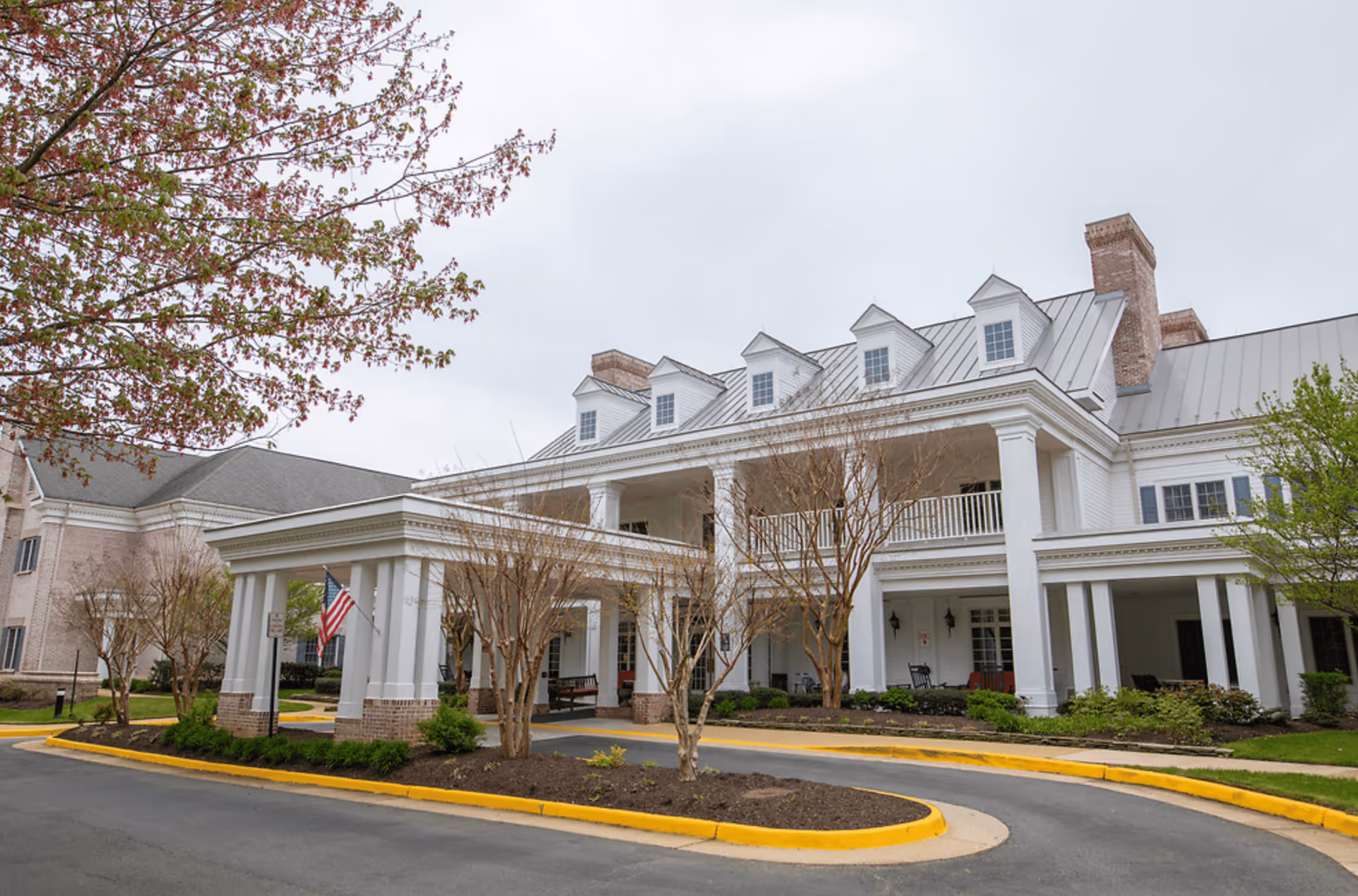 Front exterior view of a large white senior living facility building with a covered entrance, multiple windows, and dormer windows on the roof. There are leafless trees and some greenery around the building, and an American flag is displayed near the entrance.