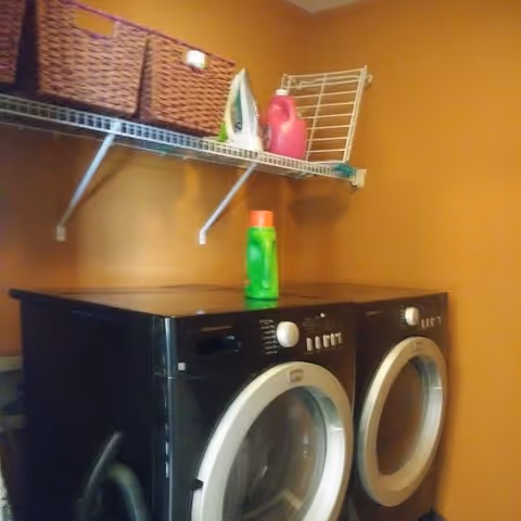 Laundry room with a black front-loading washing machine and dryer side by side against a mustard yellow wall. Above them is a white wire shelf holding two woven baskets, an iron, a pink detergent bottle, and a drying rack. A green detergent bottle is placed on top of the washing machine.