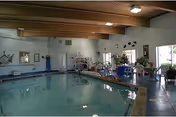Indoor swimming pool area with clear water, surrounded by chairs and tables. The room has large windows allowing natural light, plants near the windows, and a high ceiling with wooden beams.