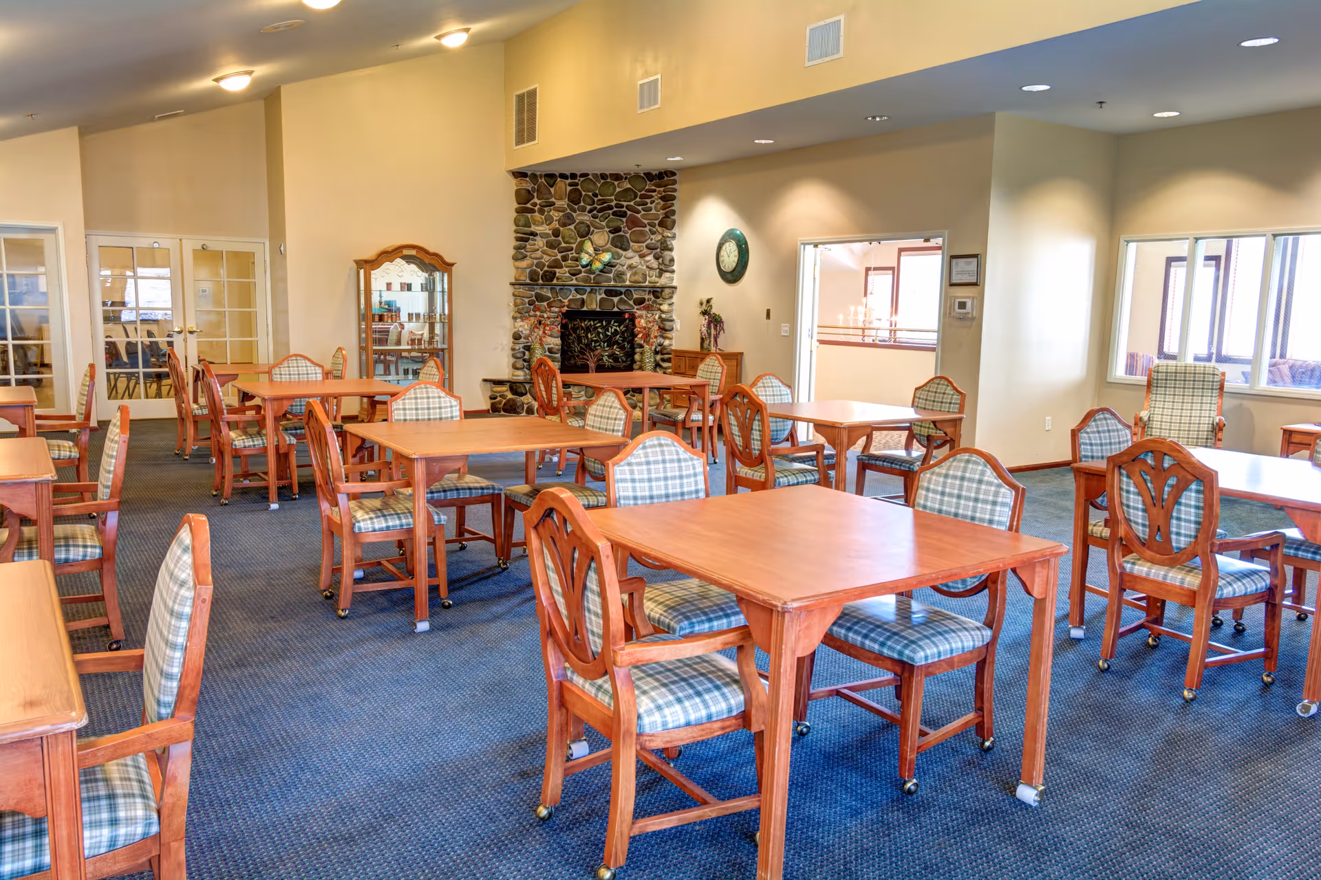 A spacious dining room with multiple wooden tables and chairs featuring plaid cushions. The room has a blue carpet, beige walls, and a stone fireplace with a clock above it. There are large windows letting in natural light and a glass cabinet in the corner.