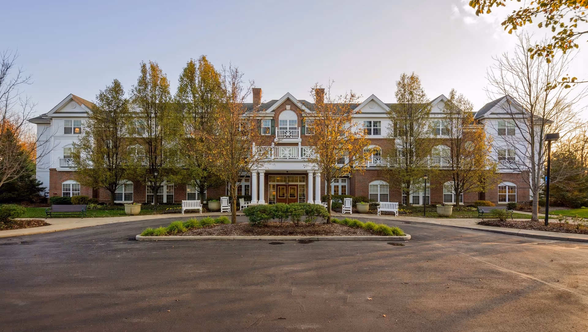 Front exterior of a three-story brick-and-white senior living building with a portico entrance, benches, and trees along a circular driveway.