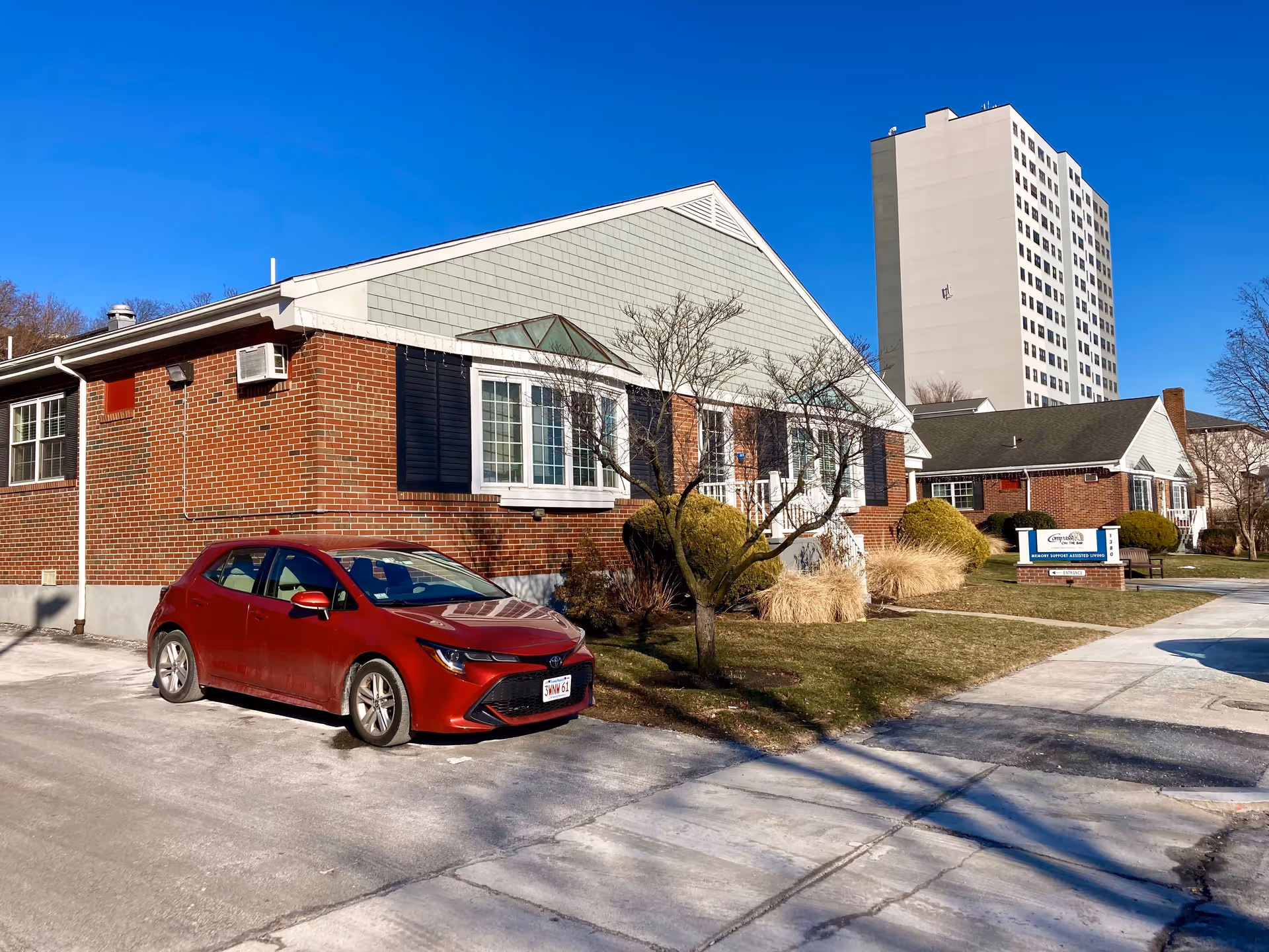 Exterior view of Compass on the Bay Memory Care Assisted Living facility showing a brick building with green siding on the upper part, a red car parked in front, a small tree, bushes, and a sign indicating the facility name and entrance. A tall white apartment building is visible in the background under a clear blue sky.