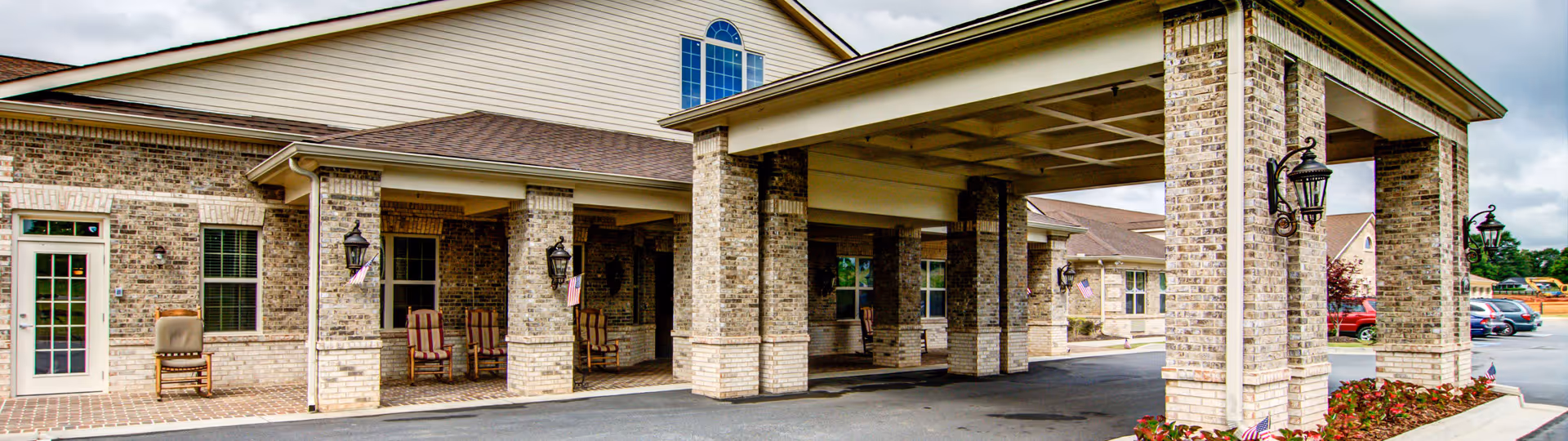 Exterior view of a senior living facility entrance with a covered drop-off area supported by brick columns. There are rocking chairs placed along the wall under the covered area, and a parking lot with cars is visible in the background. The building has beige siding and brick accents with several windows and outdoor lantern-style lights mounted on the columns.