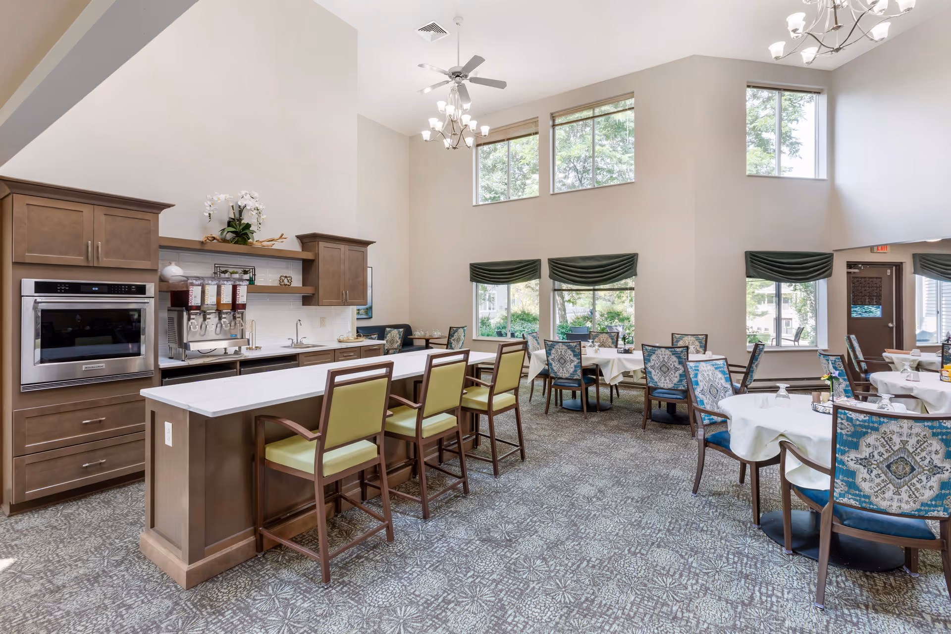 A bright dining room in a senior living facility featuring a long counter with green cushioned chairs, several round tables with white tablecloths and patterned chairs, large windows with green valances, and a high ceiling with ceiling fans and chandeliers.