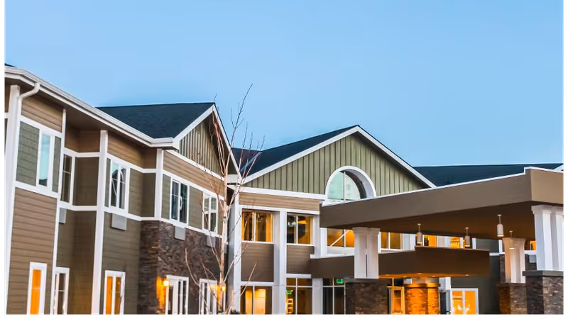 Exterior front facade and covered entrance of a multi-story senior living building with lit windows at dusk.