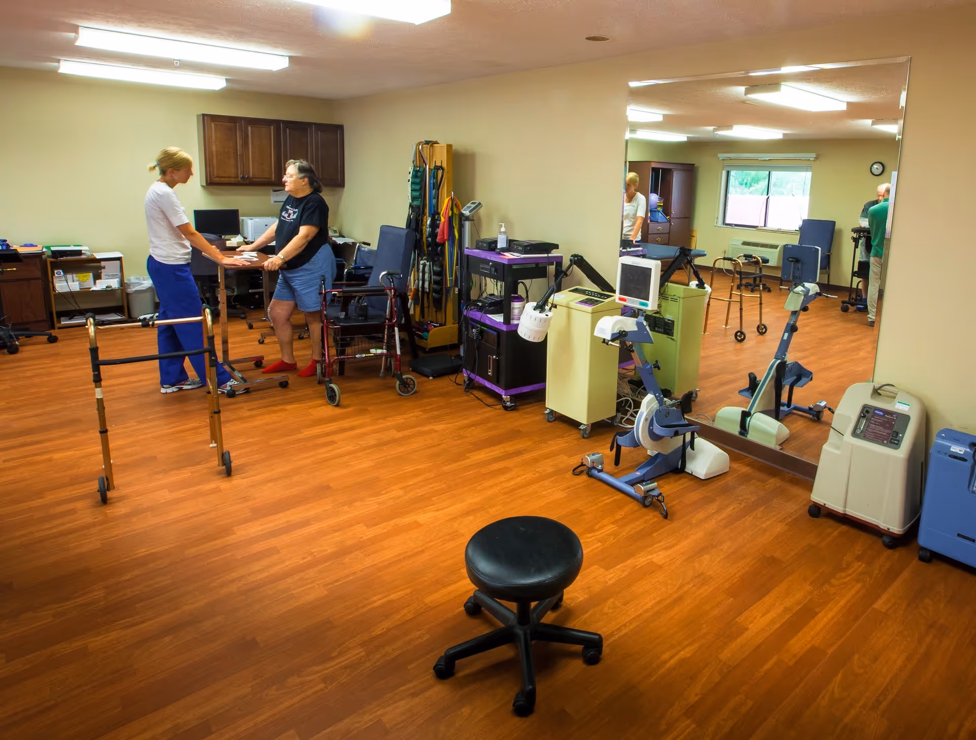 A therapy or rehabilitation room with wooden flooring, exercise equipment, walkers, and a large mirror on one wall. Two women are interacting near the center of the room, one standing and the other using a walker. The room has cabinets, a desk with a computer, and various medical or therapy devices.
