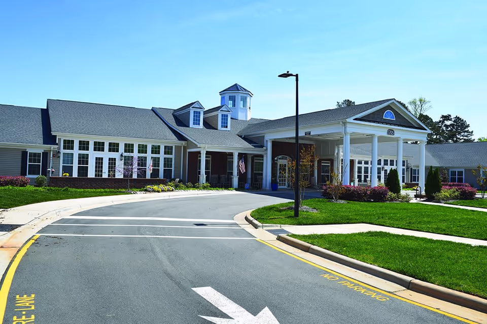 Front exterior view of a senior living facility building with a covered entrance supported by white columns, a driveway with a white arrow and yellow 'No Parking' markings, well-maintained green lawns, shrubs, and a clear blue sky.