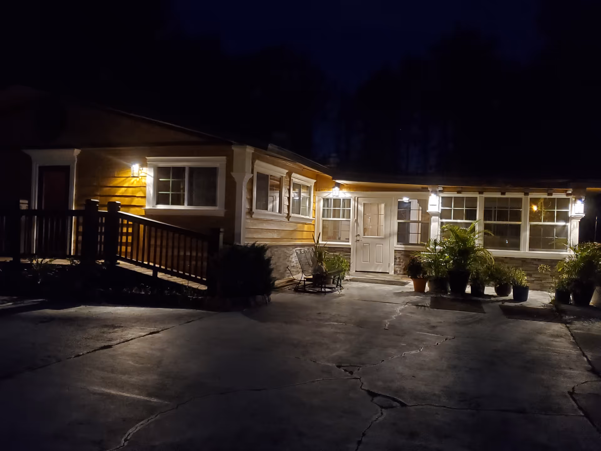 Nighttime exterior view of a single-story building with warm yellow lighting illuminating the entrance area. The building has multiple windows, a white door, potted plants near the entrance, and a ramp with railings on the left side.