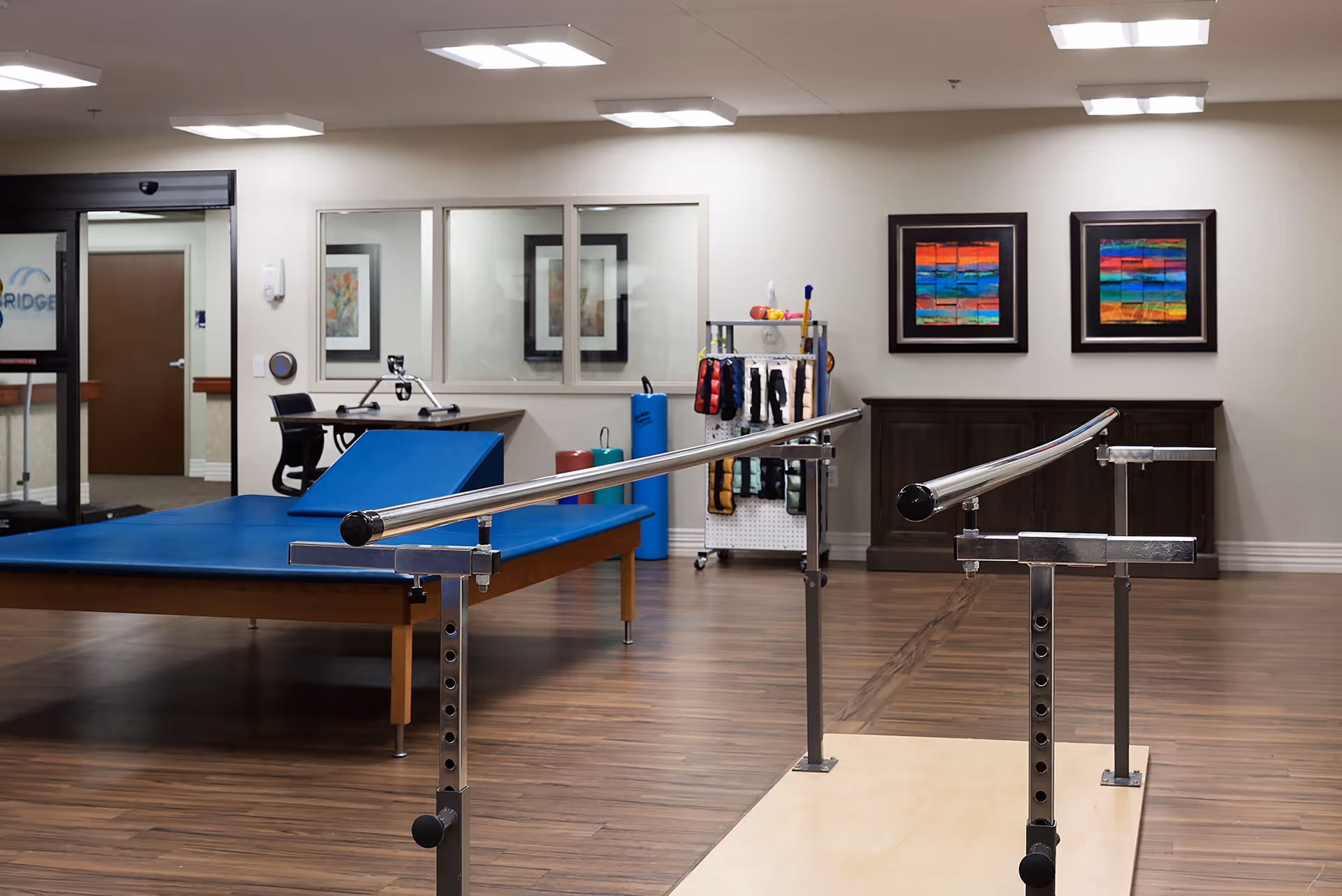 Physical therapy room with parallel bars for walking exercises, a large blue therapy table, exercise equipment, and colorful abstract artwork on the walls.