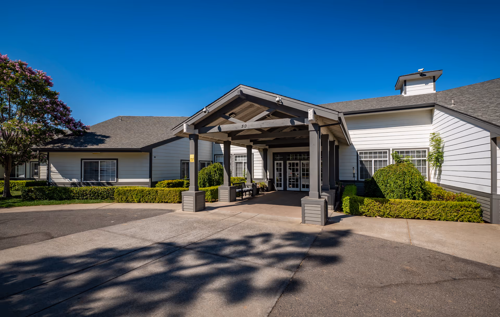 Exterior front entrance of a single-story building with a covered driveway and well-maintained landscaping including bushes and a tree with purple flowers under a clear blue sky.