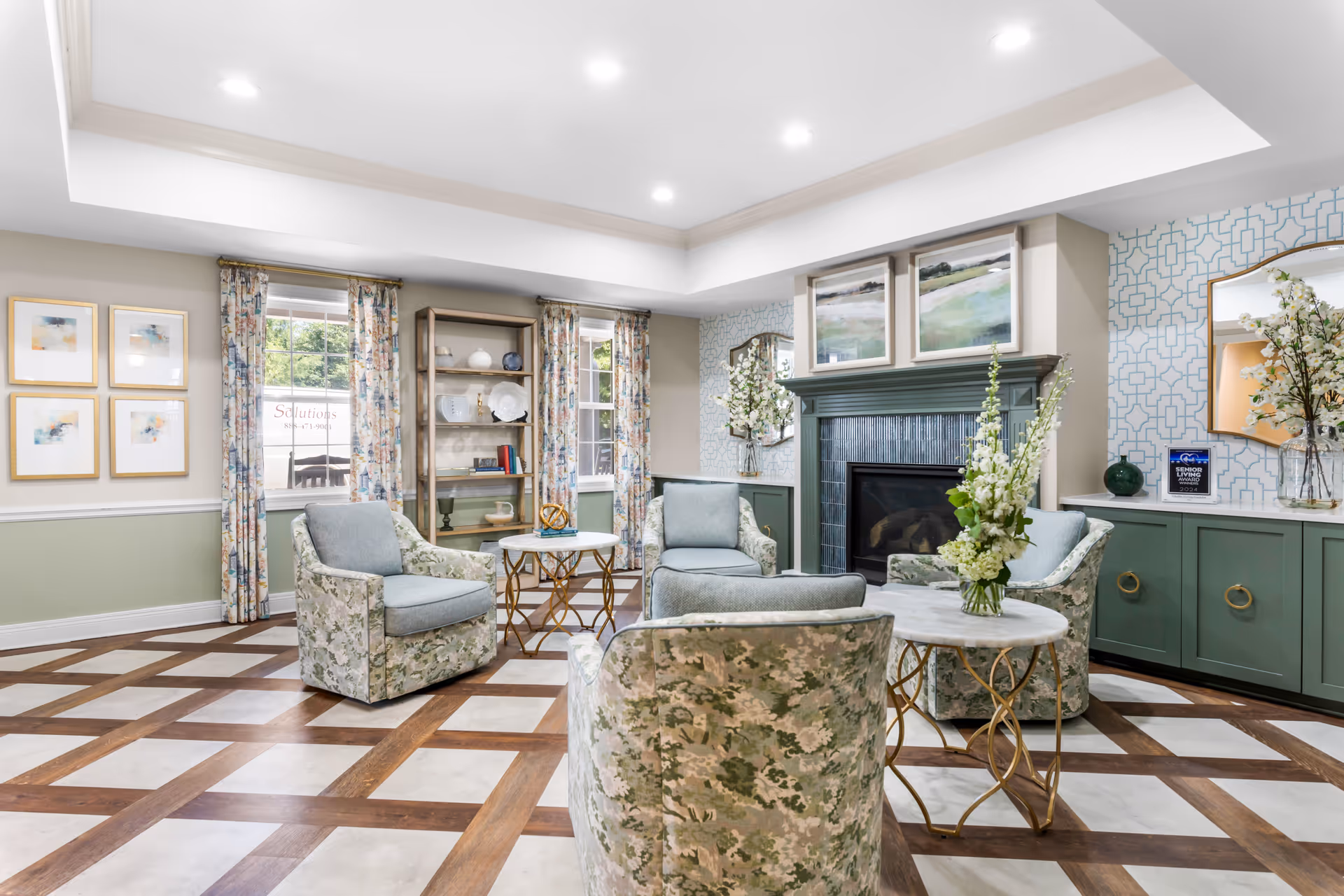 A bright and elegant living room with a patterned floor, four upholstered armchairs arranged around two small round tables with gold bases, a green fireplace with artwork above it, built-in green cabinetry with decorative items, floral curtains on windows, and soft overhead lighting.