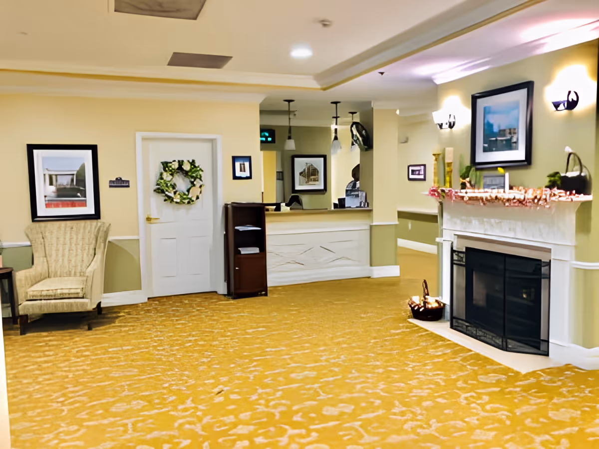Interior view of a senior living facility lobby area with a yellow patterned carpet, a beige armchair, a white door decorated with a green wreath, a reception desk with a mirror behind it, and a fireplace adorned with candles and decorations.