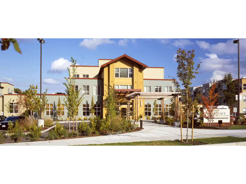 Exterior view of Park Place assisted living facility with a modern multi-story building, landscaped garden with young trees and shrubs, a curved driveway, and a clear blue sky with some clouds.