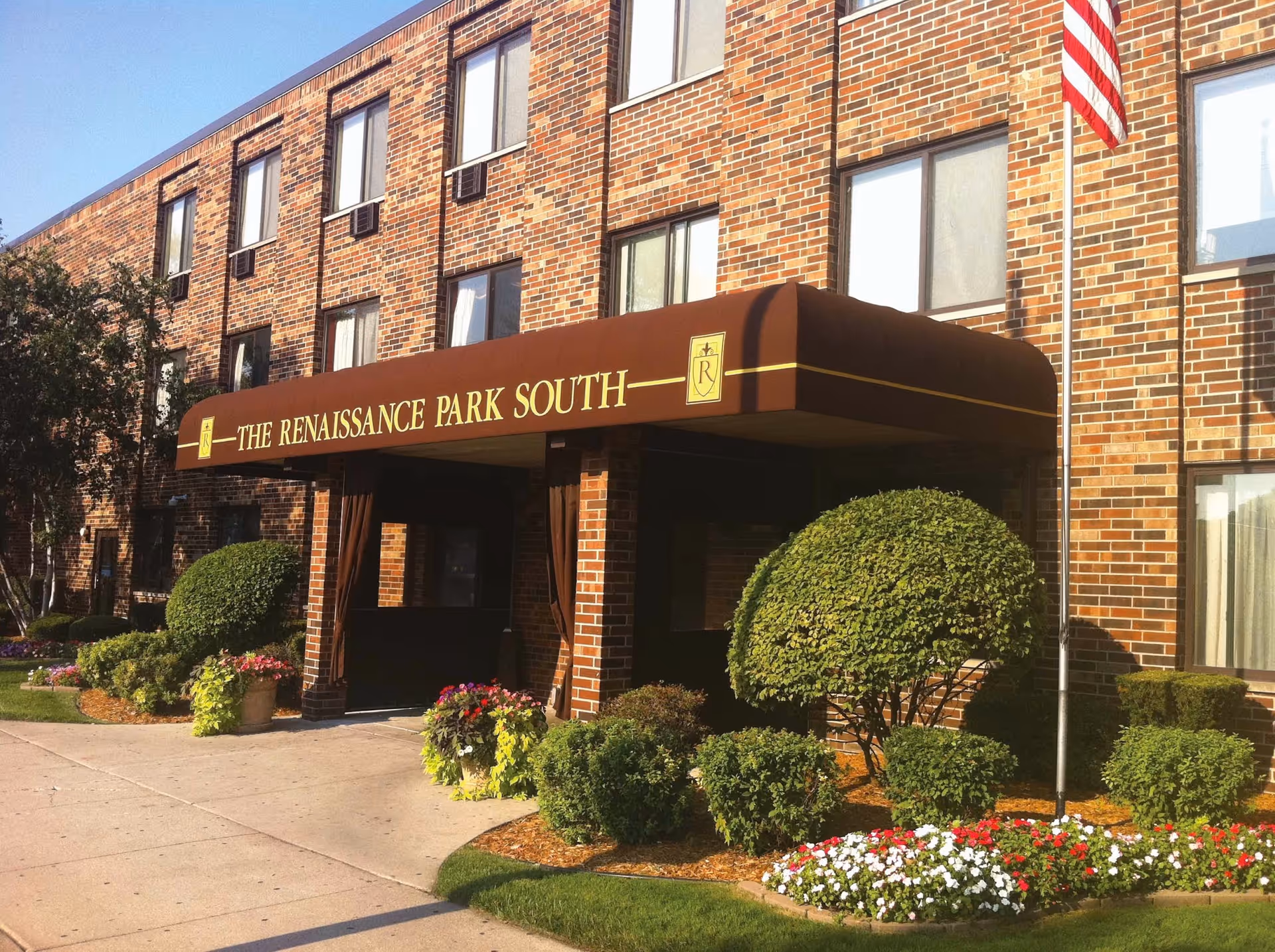 Exterior view of a brick building with a brown awning that reads 'THE RENAISSANCE PARK SOUTH'. The entrance is surrounded by neatly trimmed bushes and colorful flower beds. An American flag is visible on the right side of the image.