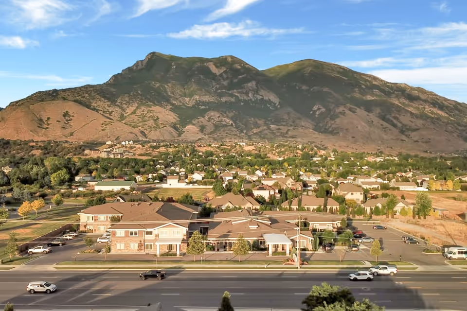 Aerial view of a senior living facility named Highland Glen with a large parking lot and multiple buildings, set against a backdrop of a mountain range and a suburban neighborhood under a partly cloudy sky.