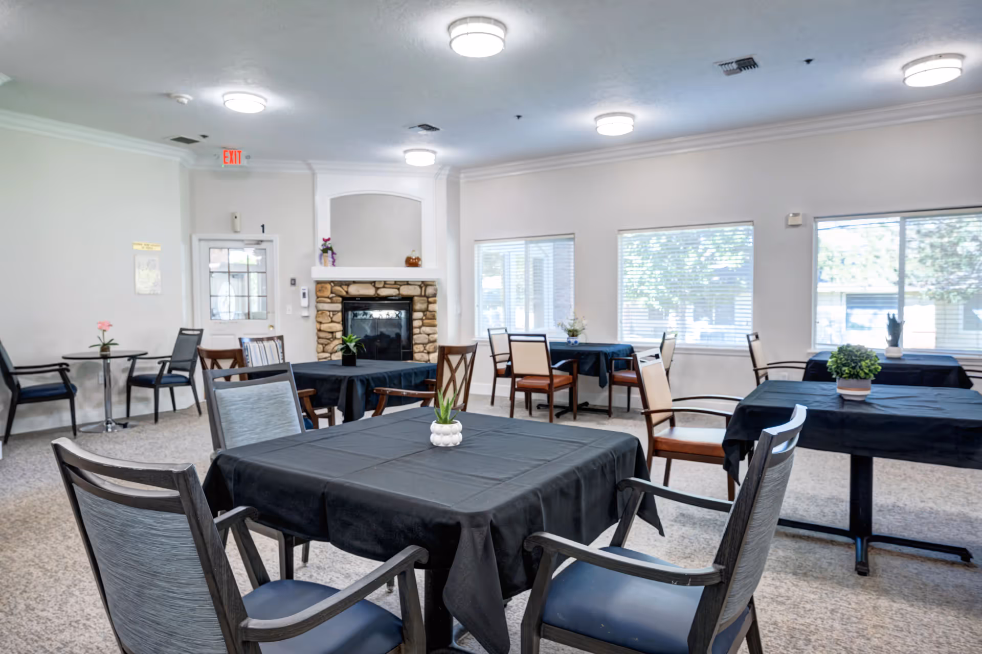 Bright communal dining room with multiple tables covered in black tablecloths, chairs, and a stone fireplace.