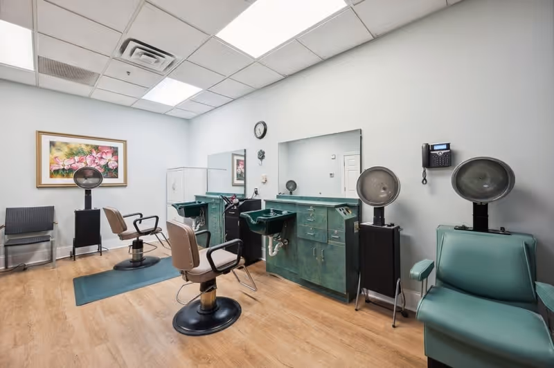 Interior of a salon room with two salon chairs facing mirrors and sinks, a green salon dryer chair with two hooded hair dryers, a wall-mounted phone, a framed floral painting, and a clock on the wall. The room has light-colored walls and wood flooring.