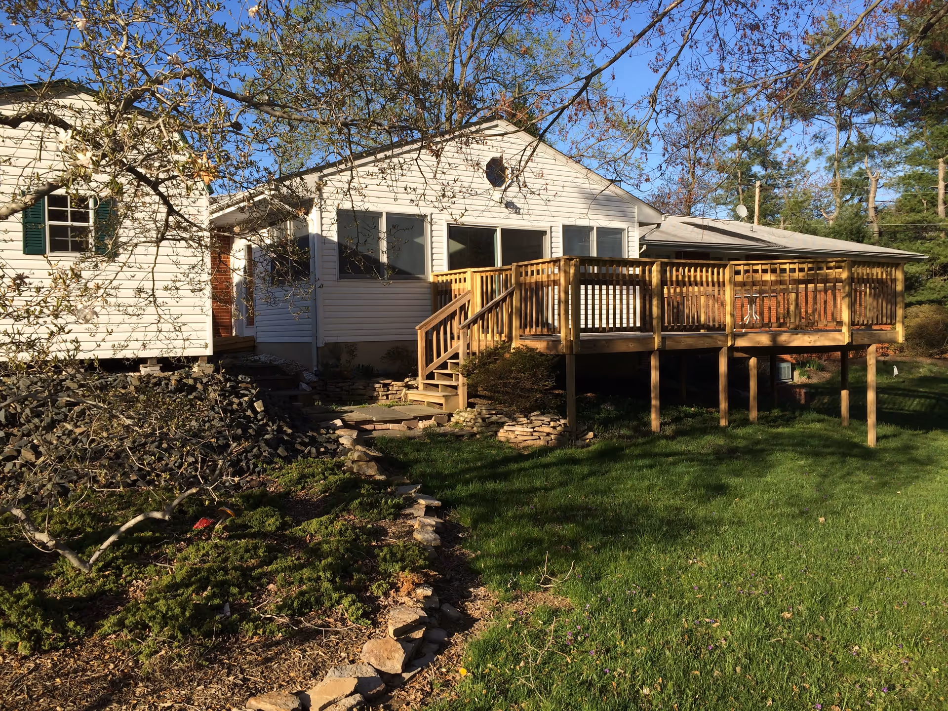 Exterior view of a single-story white house with a wooden deck and stairs leading down to a grassy yard. There are trees and shrubs around the house, and the sky is clear and blue.
