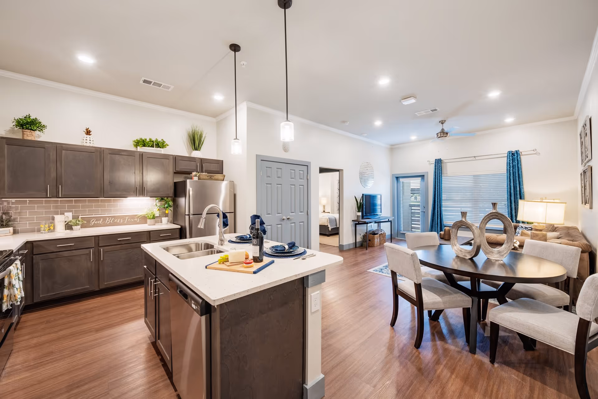 A modern open-concept interior of a senior living facility showing a kitchen with dark cabinets, stainless steel appliances, and a white island with a sink and place settings. Adjacent to the kitchen is a dining area with a dark oval table and six upholstered chairs. In the background, there is a living room area with a sofa, a TV on a stand, blue curtains, and a ceiling fan. A bedroom is visible through an open doorway.