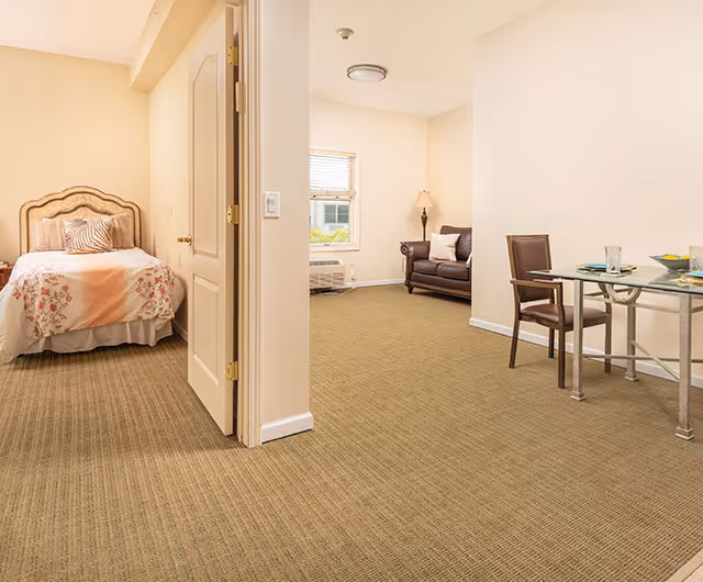 Interior view of a senior living facility room at Lakewood Courtyard showing a bedroom with a bed and floral bedding on the left, and an adjacent living area with a brown armchair, a floor lamp, and a small glass dining table with two chairs on the right. The room has beige walls and carpeted flooring.